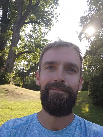 Selfie von Mann mit Bart. Stefan Reitinger sein Name. Im Hintergrund sind hohe Bäume, durch die die Sonne blinzelt.