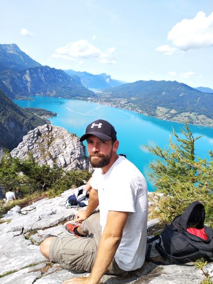 Mann mit Kappe sitzt auf einem Felsen. Im Hintergrund liegt der türkis blaue Attersee.